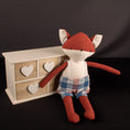 Charger l'image dans la visionneuse de la galerie, Un adorable renard en tissus couleur rouille et écru qui porte un short à bretelle en tissu à carreaux assorti. Il est adossé à un petit meuble en bois.